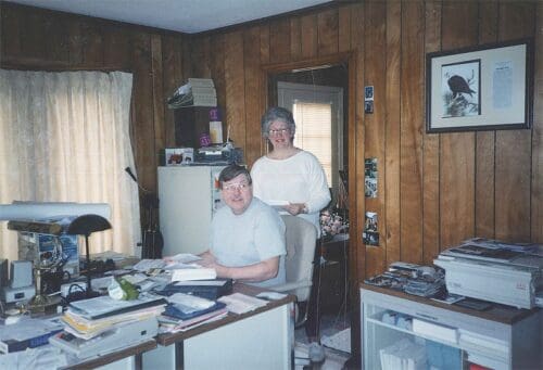 A man and woman smiling for the camera in an old room with may papers and files around